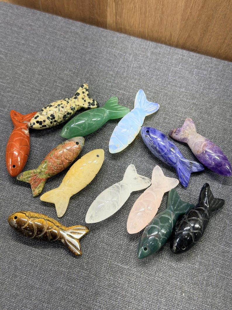 May include: A selection of carved stone fish in diverse colours and patterns. The fish include orange, green, yellow, white, pink, blue, and black. Some have speckled patterns, while others are solid colours. The fish are arranged on a grey surface.