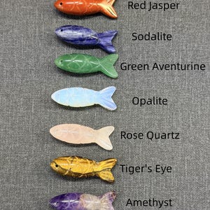 May include: Seven gemstone fish figurines in a variety of colours. The fish are carved from different gemstones, including red jasper, sodalite, green aventurine, opalite, rose quartz, tiger's eye, and amethyst. Each fish is labelled with its gemstone type. The fish are displayed on a grey surface.