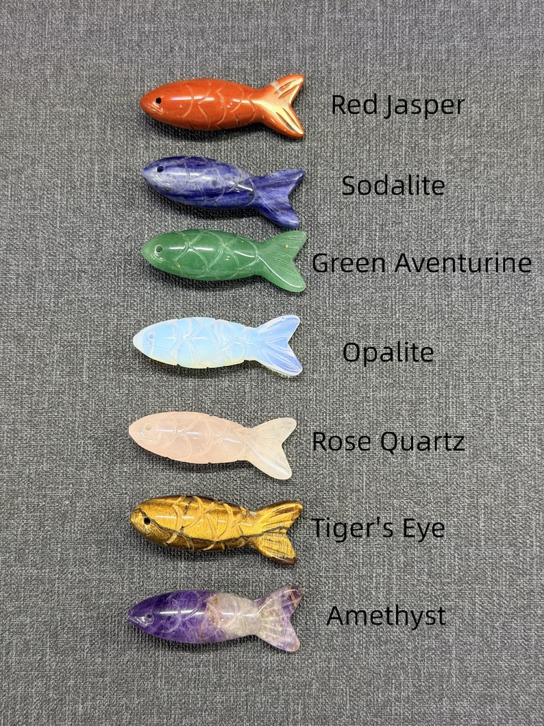 May include: Seven gemstone fish figurines in a variety of colours. The fish are carved from different gemstones, including red jasper, sodalite, green aventurine, opalite, rose quartz, tiger's eye, and amethyst. Each fish is labelled with its gemstone type. The fish are displayed on a grey surface.