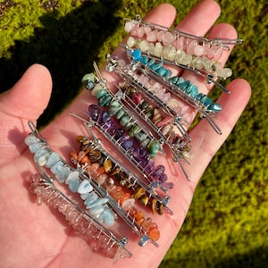 May include: A collection of gemstone hair clips in various colors, including pink, blue, green, and orange. Each clip features a silver metal base and is adorned with small, irregularly shaped stones. The clips are displayed on a hand against a green, mossy background.