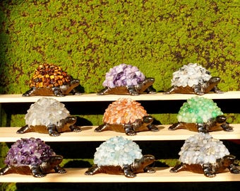May include: A collection of decorative turtle figurines with gemstone shells. The turtles have bronze-colored bodies and are adorned with various colored gemstones, including amber, amethyst, quartz, and jade. They are displayed on wooden shelves against a green moss background.