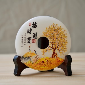 Könnte beinhalten: Weiße runde Keramikplatte mit einem Loch in der Mitte. Die Platte hat ein gemaltes Design mit einem Baum mit gelben Blättern, zwei Kranichen und chinesischen Schriftzeichen. Die Platte steht auf einem Holzständer.