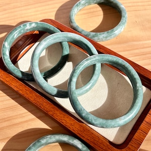 May include: A collection of green jade bangles, each with a circular design, are presented on a wooden tray. The bangles exhibit a smooth, polished finish and different shades of green. The tray has a dark brown frame and a light beige interior.