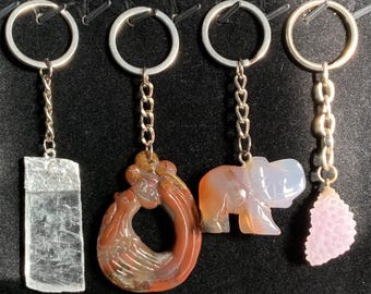 May include: Four keychains with different charms. The charms are a clear crystal, a red and brown swirl, a pink and white elephant, and a pink crystal. The keychains are all silver.