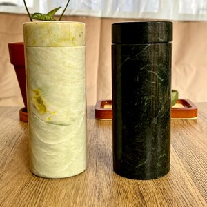 May include: Two cylindrical stone containers. One is light green with yellow and white veining, the other is black with green veining. Both have a matching lid. They are displayed on a wooden surface.