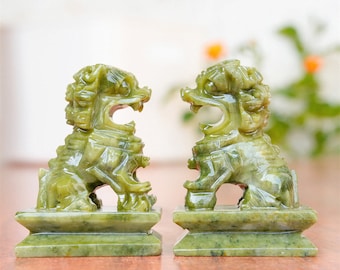 May include: A pair of green jade foo dog statues. The statues are facing each other and are sitting on square bases.