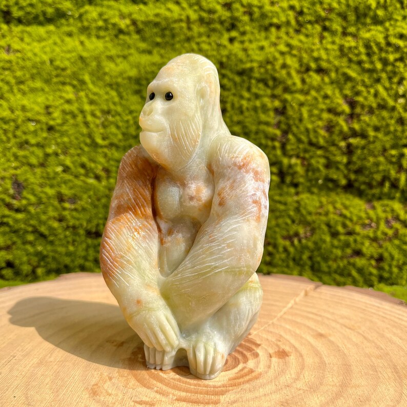 May include: A hand-carved stone sculpture of a gorilla sitting with its arms crossed. The sculpture is made of a light green stone with brown and orange markings.