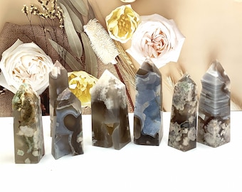 May include: A collection of seven agate crystal towers in varying shades of gray, white, and blue. Each tower has a unique pattern and shape, with a polished, smooth surface. The crystals are arranged on a white surface, with floral decorations in the background.