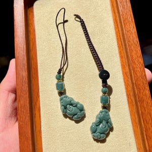 Puede incluir: Dos colgantes de jade con intrincadas tallas de criaturas míticas. Cada colgante es de color verde apagado y está ensartado en un cordón marrón con cuentas decorativas. Los colgantes se exhiben en una bandeja de madera.