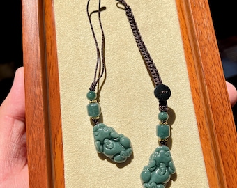 May include: Two jade pendants with intricate carvings of mythical creatures. Each pendant is a muted green color and is strung on a brown cord with decorative beads. The pendants are displayed on a wooden tray.
