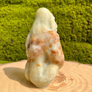 May include: A hand-carved green and orange stone sculpture of a person sitting with their legs crossed. The sculpture is made of a type of stone that has a mottled appearance.