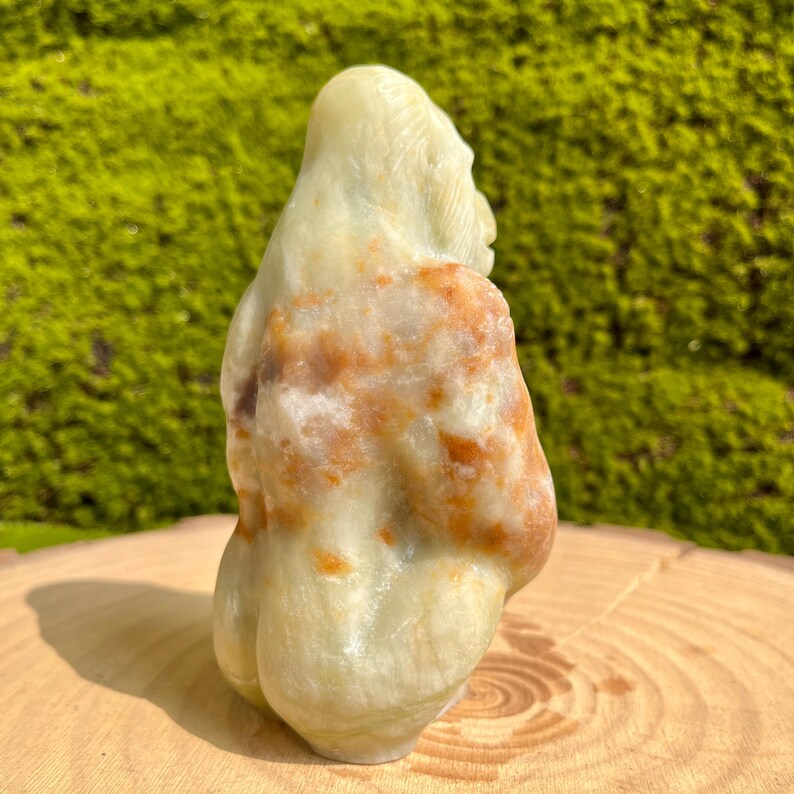 May include: A hand-carved green and orange stone sculpture of a person sitting with their legs crossed. The sculpture is made of a type of stone that has a mottled appearance.