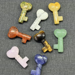 Op de afbeelding: Een verzameling van acht edelstenen sleutels met hartvormige koppen. De sleutels zijn in verschillende kleuren, waaronder groen, wit, geel, rood, bruin, roze en blauw. Elke sleutel heeft een uniek patroon en textuur, tegen een grijze achtergrond.