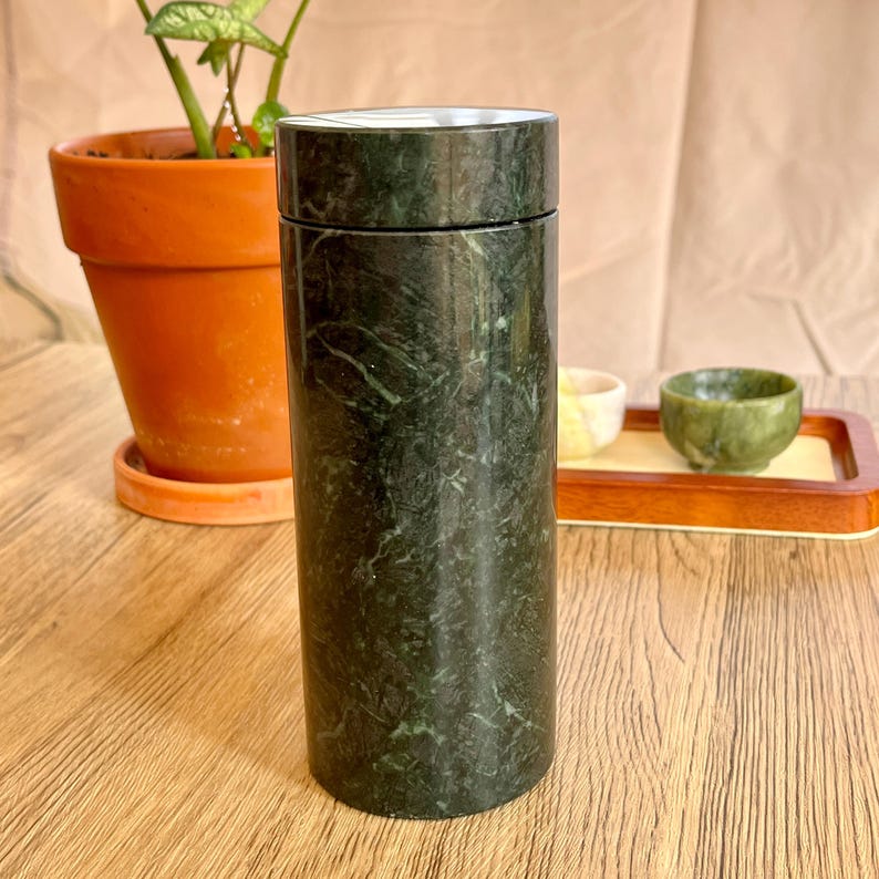May include: A cylindrical, dark green marble tea container with a matching lid. The container is displayed on a wooden surface, with a terracotta pot and small tea cups in the background. The marble has a natural, veined pattern.