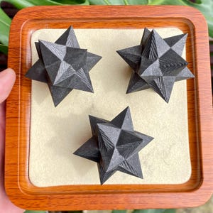 May include: Three dark gray, star-shaped decorative objects with textured surfaces are displayed on a beige surface within a wooden tray. The objects have multiple points and a geometric design, creating a striking visual effect.