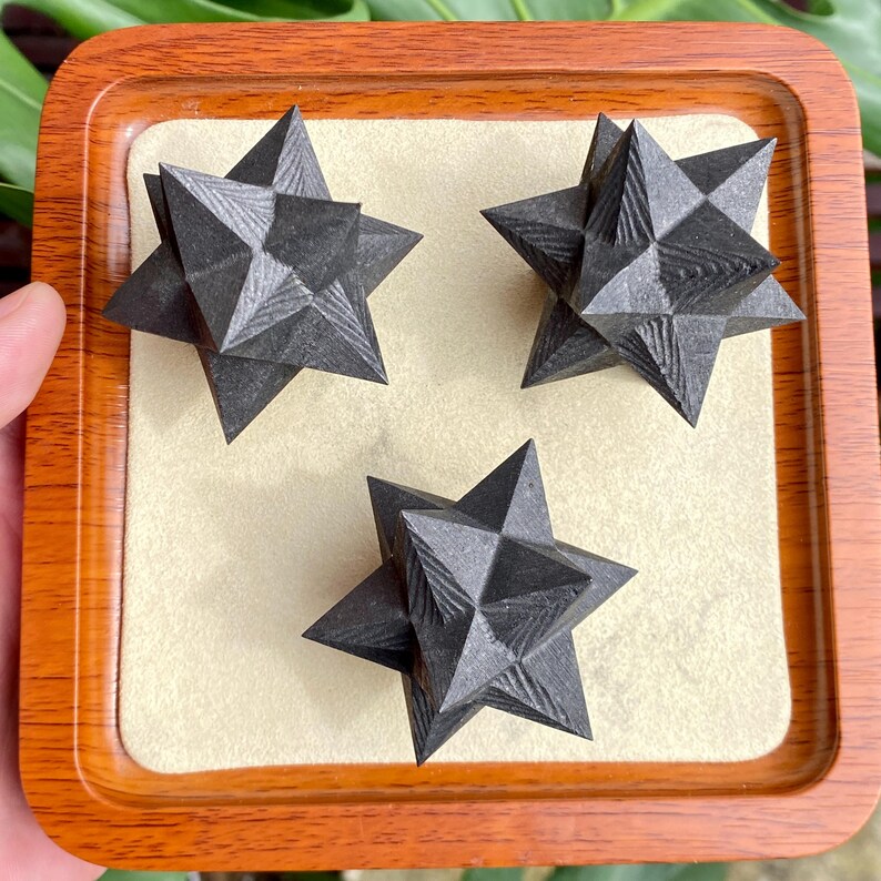 May include: Three dark gray, star-shaped decorative objects with textured surfaces are displayed on a beige surface within a wooden tray. The objects have multiple points and a geometric design, creating a striking visual effect.