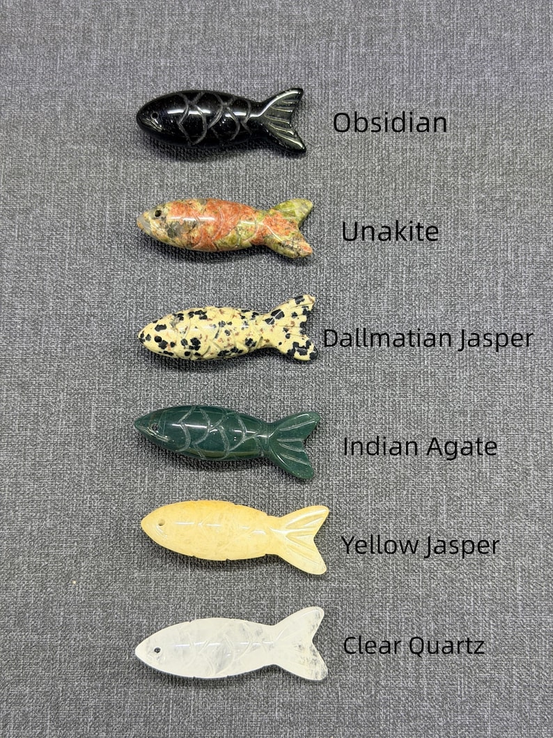 May include: Six carved stone fish figurines in a row. The fish are made from different stones, including obsidian and unakite. Each fish has a unique colour and pattern, with the names of the stones labelled next to each fish.