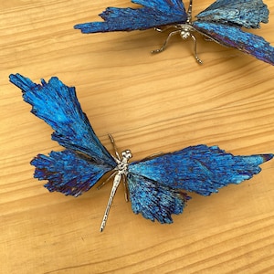 May include: Two decorative dragonfly sculptures with textured blue wings and silver-colored bodies. The wings have a layered, crystalline appearance with hints of purple. The dragonflies are set against a light brown wooden surface.