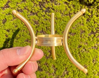 May include: A gold-colored metal candle holder with a unique design. The holder features curved arms and a central ring, with a small symbol etched on the ring. The holder is held against a green, mossy background.