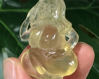 May include: A translucent yellow figurine of a seated woman with long flowing hair. The sculpture has a rounded form and is carved with delicate details. The figure is holding her hands near her chest. The background is blurred green.