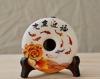 May include: A white ceramic donut-shaped ornament with a hole in the center. The ornament is decorated with a colorful design of koi fish and Chinese characters that read "Nine Fish Bring Prosperity". The ornament is displayed on a small wooden stand.