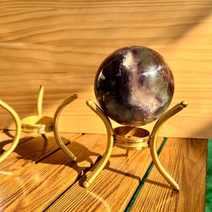 Puede incluir: Una bola de cristal esférica pulida con tonos morados y blancos, exhibida sobre un soporte de metal dorado. El soporte tiene un diseño curvo y una base circular. El fondo es una superficie de madera.
