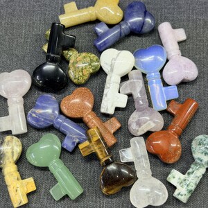 Op de afbeelding: Een verzameling edelstenen sleutels in verschillende kleuren, waaronder geel, blauw, groen en bruin. De sleutels hebben hartvormige sleutelkoppen en zijn gerangschikt op een grijze ondergrond. De edelstenen hebben verschillende patronen en texturen.
