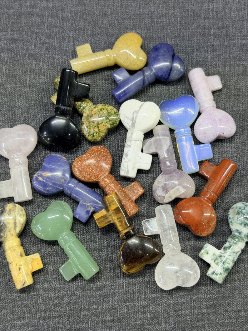 Op de afbeelding: Een verzameling edelstenen sleutels in verschillende kleuren, waaronder geel, blauw, groen en bruin. De sleutels hebben hartvormige sleutelkoppen en zijn gerangschikt op een grijze ondergrond. De edelstenen hebben verschillende patronen en texturen.