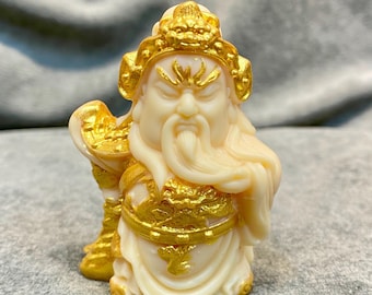 May include: Ivory-colored figurine of a seated deity with a long white beard and flowing hair. The figurine is adorned with gold-colored accents on the crown, clothing, and the object held in the left hand. The figure has a stern expression.