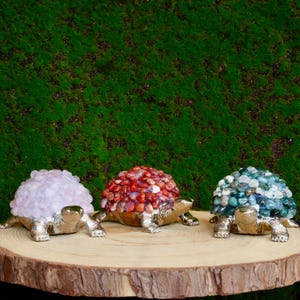 Peut inclure: Trois figurines de tortues décoratives avec des carapaces en pierres précieuses. L'une a une carapace rose clair, une autre rouge et la troisième bleue et verte. Les tortues ont des accents dorés et argentés, exposées sur une surface en bois.