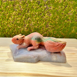 Puede incluir: Una escultura detallada de una ardilla en tonos naranja, rosa y verde, descansando sobre una base de piedra gris. La ardilla tiene una cola tupida y está posada de forma realista. La escultura está sobre un fondo verde borroso.