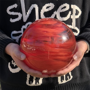 Può includere: Un grande globo di vetro rosso con motivi a vortice. Il globo è tenuto con entrambe le mani e la luce si riflette sulla superficie liscia. Lo sfondo è un maglione nero con scritte bianche.