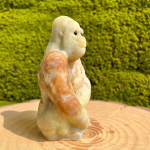 May include: A hand-carved stone sculpture of a gorilla, sitting with its arms crossed. The sculpture is made of a light green and brown stone with white accents. The gorilla's eyes are black and the sculpture is detailed with realistic features.
