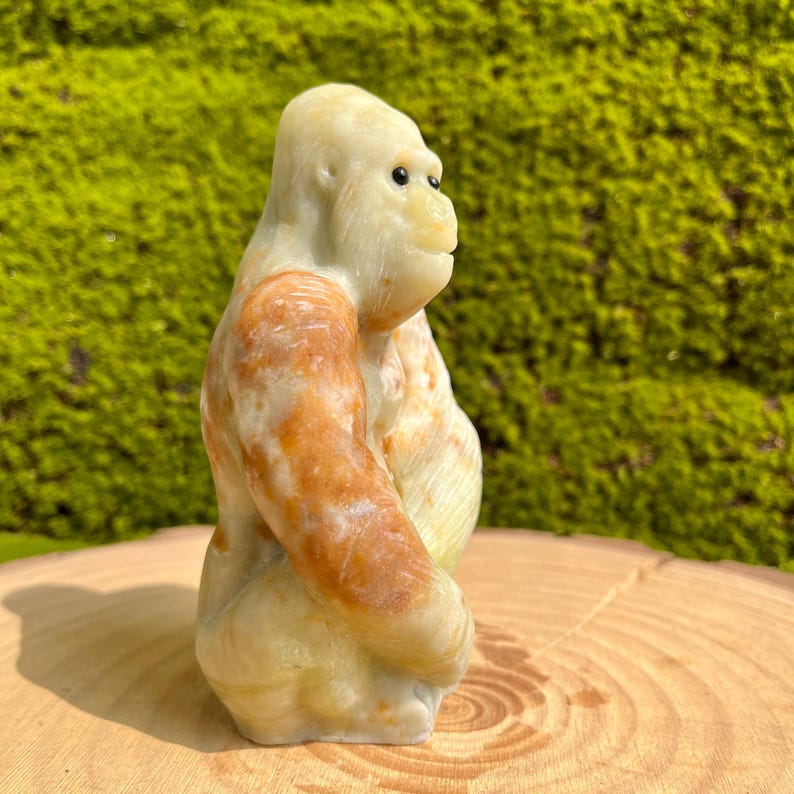 May include: A hand-carved stone sculpture of a gorilla, sitting with its arms crossed. The sculpture is made of a light green and brown stone with white accents. The gorilla's eyes are black and the sculpture is detailed with realistic features.