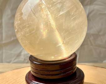 May include: A large, clear, yellow citrine crystal sphere on a dark wood stand. The sphere has a rainbow iridescence.