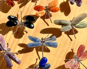May include: A collection of gemstone dragonfly figurines in various colors, including red, orange, black, blue, purple, and green. Each dragonfly has silver-colored metal wings and body. The gemstones are polished and have a smooth texture.