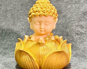 May include: A small, golden Buddha statue seated in a lotus flower. The Buddha figure has closed eyes and a serene expression. The lotus petals are detailed and painted in a metallic gold finish. The statue is a decorative item.