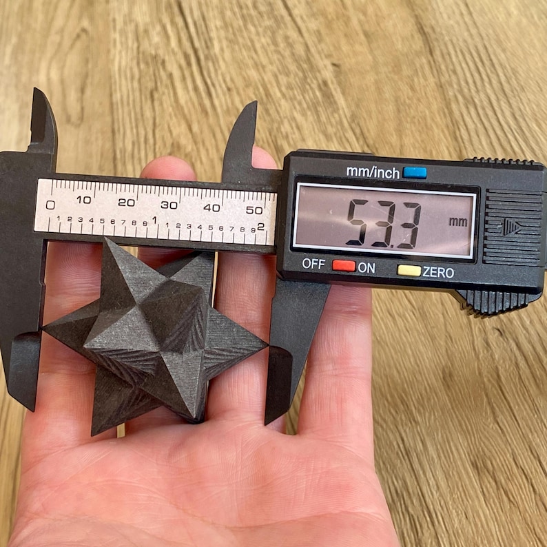 May include: A dark gray, geometric star-shaped object is measured with a digital caliper. The caliper displays 53.3 mm, approximately 2.1 inches. The object has a textured surface and is held against a light brown wooden background.