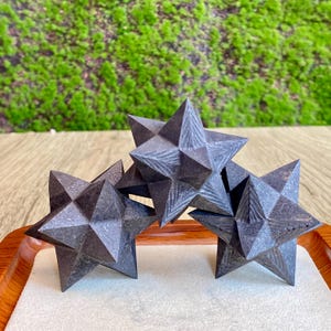 May include: Three dark gray, multi-faceted star-shaped decorative objects. The stars are arranged on a white surface within a wooden tray, with a green moss background. The objects have a geometric design and are likely made of stone or a similar material.