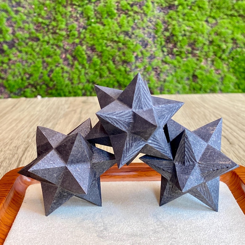 May include: Three dark gray, multi-faceted star-shaped decorative objects. The stars are arranged on a white surface within a wooden tray, with a green moss background. The objects have a geometric design and are likely made of stone or a similar material.