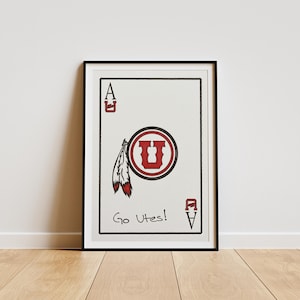 May include: Framed art print featuring a playing card design. The card is white with a red and black University of Utah logo and feather graphic. The text "Go Utes!" is written at the bottom. The card is framed in a black frame.