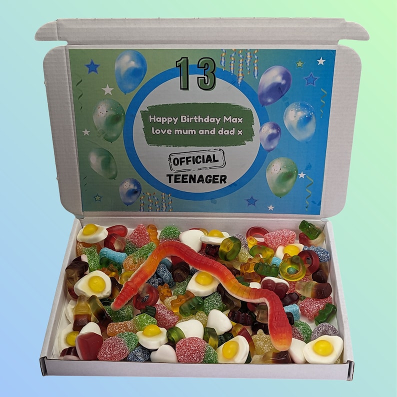 Op de afbeelding: Een open witte doos gevuld met een verscheidenheid aan kleurrijke gummy snoepjes. Het deksel van de doos heeft een blauw en groen ontwerp met ballonnen en de tekst "13", "Happy Birthday Max love mum and dad x" en "OFFICIAL TEENAGER".