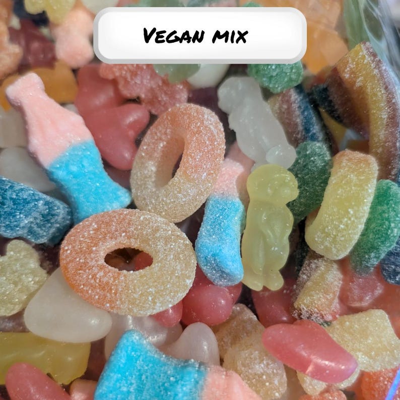 Op de afbeelding: Een close-up van een mix van kleurrijke veganistische snoepjes. De snoepjes omvatten ringvormige, flesvormige en gummybeer-vormige snoepjes in verschillende kleuren zoals roze, oranje, blauw en geel. De tekst "VEGAN MIX" wordt weergegeven in een witte doos.
