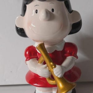 May include: A ceramic figurine of a cartoon character, wearing a red dress with white trim and holding a gold-colored trumpet. The figurine has black hair and brown shoes. The character is smiling.