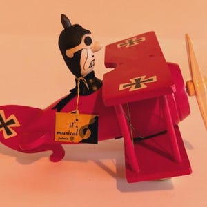 May include: A red biplane toy with a cartoon pilot figure. The plane has a yellow propeller and black and white accents. The wings feature black cross symbols. A tag reads "it's musical".