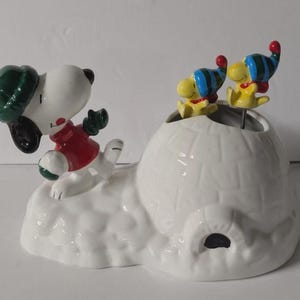 May include: A ceramic Snoopy figurine in a winter scene. Snoopy is wearing a red jacket, green hat, and gloves, and is positioned on a snow-covered base. Two Woodstock figures with blue and yellow striped hats are perched atop a white igloo.