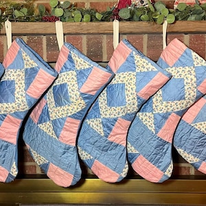 May include: Six quilted Christmas stockings featuring a patchwork design of light blue, soft pink, and a cream-colored fabric with a small floral pattern. Each stocking is constructed with a geometric arrangement of these fabric squares and triangles.