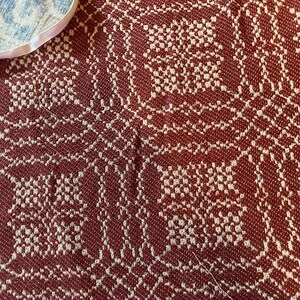 May include: A woven textile featuring a deep red and off-white geometric pattern. The fabric displays a dense, repeating grid-like design with intricate interlocking shapes. A small portion of a white circular edge is visible in the upper corner.