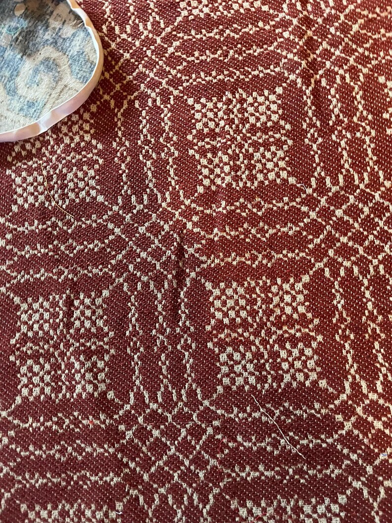 May include: A woven textile featuring a deep red and off-white geometric pattern. The fabric displays a dense, repeating grid-like design with intricate interlocking shapes. A small portion of a white circular edge is visible in the upper corner.