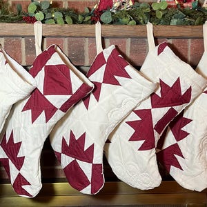May include: Five quilted Christmas stockings hanging in a row, each featuring a white base with a deep red geometric star pattern. The stockings are constructed with visible stitching and fabric loops at the top for hanging on a fireplace mantel.